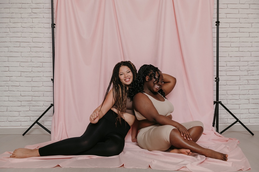 Two joyful plus size African women in sportswear sitting and bonding on pink fabric background together